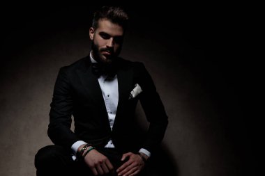 Siyah smokin giyen genç işadamı karanlık stüdyo geçmişine bakarak kameraya bakıyor.
