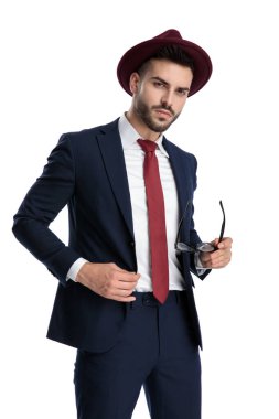 Yakışıklı iş adamı bordo şapka takıyor ve elinde gözlük tutarken beyaz stüdyo arka planında ciddi bir ceketi tamir ediyor.