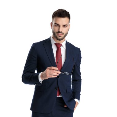 Genç işadamı, donanma takımı giymiş, el ele tutuşmuş, gözlük tutuyor ve beyaz stüdyo arka planındaki kameraya ciddi ciddi bakıyor.