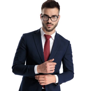Genç iş adamı ayakta durup kollarını düzeltirken beyaz stüdyo arka planında ciddi bir tavır sergiliyor.