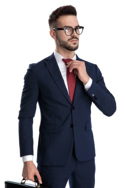 Gözlüklü çekici bir iş adamı beyaz stüdyo arka planında kravatını sert bir tavırla düzeltirken elinde evrak çantası tutuyor.