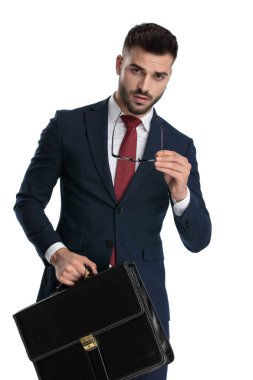 Çekici iş adamı donanma takımı giyiyor ve evrak çantası tutuyor. Beyaz stüdyo arka planında duruşu olan gözlükleri çıkartıyor.