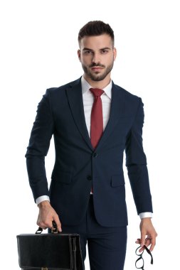 Yakışıklı iş adamı donanma takımı giyiyor ve bir elinde evrak çantası, diğer elinde gözlüklerle beyaz stüdyo arka planında ciddi bir şekilde duruyor.