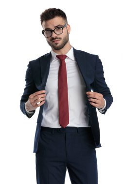 Gözlüklü çekici bir iş adamı beyaz stüdyo arka planında havalı bir tavırla poz verirken ceketini düzenliyor.