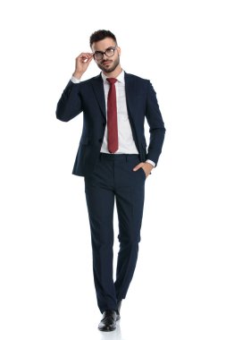 Genç işadamı donanma takımı giyiyor, cebinde yürüyor ve beyaz stüdyo arka planında gözlük takıyor.