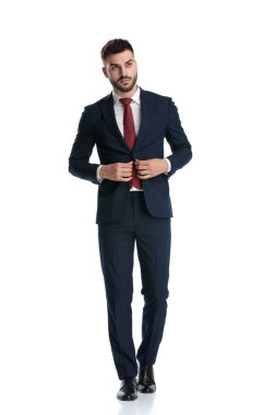 Genç işadamı ceketini açarken yürürken donanma giysisi giyiyor ve beyaz stüdyo geçmişine ciddi ciddi bakıyor.