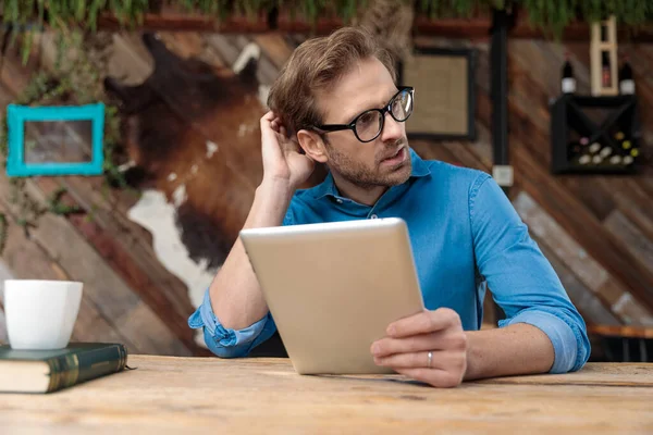 Ležérní Muž Brýlích Sedí Stolem Drží Tablet Zatímco Škrábe Hlavu — Stock fotografie