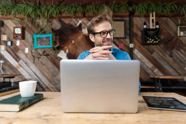机の上に眼鏡をかけた青年がノートパソコンで仕事をしてコーヒーショップで幸せそうな顔をして — ストック写真