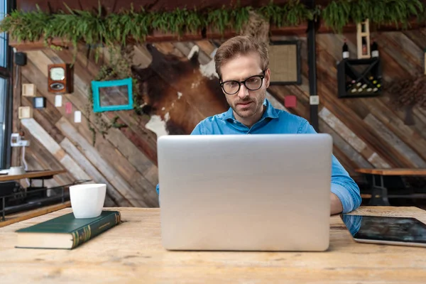 デスクに座ってコーヒーショップを中心にノートパソコンを勉強する眼鏡をかけた魅力的なビジネスマン — ストック写真