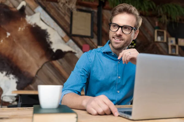 有魅力的商人 戴着眼镜 坐在办公桌前 在笔记本电脑上工作 一边高兴地看着远处的咖啡店 — 图库照片