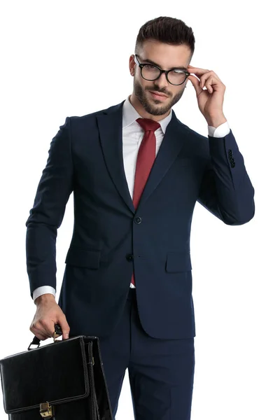 海軍のスーツを着た若い実業家が片手でブリーフケースを持ちながら白いスタジオの背景に幸せそうな眼鏡をかけながら — ストック写真