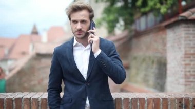 Rahat ve akıllı iş adamı, bir eli cebinde tuğla bir duvara yaslanmış telefonla konuşuyor ve endişeleniyor.