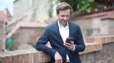 Tuğla duvara yaslanan bir iş adamı, çantasını tutarken telefonuyla kitap okuyor ve kendini eğlendiriyor.