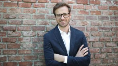 Çekici, mutlu ve gözlüklü bir işadamı elleri göğsünde bir tuğla duvarın karşısında duruyor, gülümsüyor ve kameraya bakıyor.