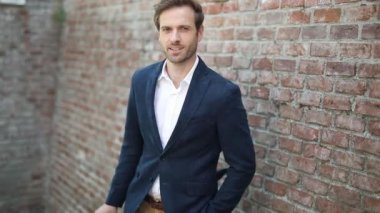 Mutlu yakışıklı iş adamı bir eli cebinde poz veriyor, gülümsüyor ve sonra gözlüğünü tuğla duvara dayıyor.