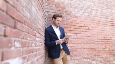 Duvara yaslanmış, donanma kıyafeti giymiş ve kameraya gülümseyen zarif, genç bir işadamının tabletini okuyuşunu yakından çek.