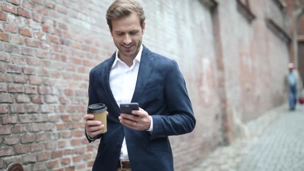セクシーなエレガントなビジネスマンが海軍のスーツを着てレンガの壁の近くの彼の携帯電話にテキストメッセージを送りながらコーヒーを持っています — ストック動画