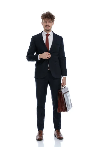 スーツを着て白いスタジオの背景に立っている間に幸せなビジネスマンは彼のジャケットをボタンを外し ブリーフケースを保持 — ストック写真
