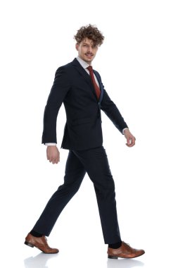 Takım elbise giyerken gülümseyen ve beyaz stüdyo arka planında yürüyen mutlu iş adamının yan görüntüsü