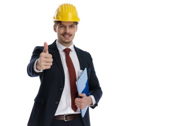 Pozitif iş adamı elinde bir pano tutarken ve miğfer ve takım elbise giyerken iyi hareket ederken beyaz stüdyo arka planında duruyor.