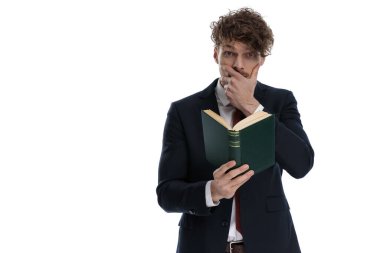 Şaşırmış iş adamı kitap okuyor ve takım elbise giyip beyaz stüdyo arka planında dikilirken ağzını kapatıyor.