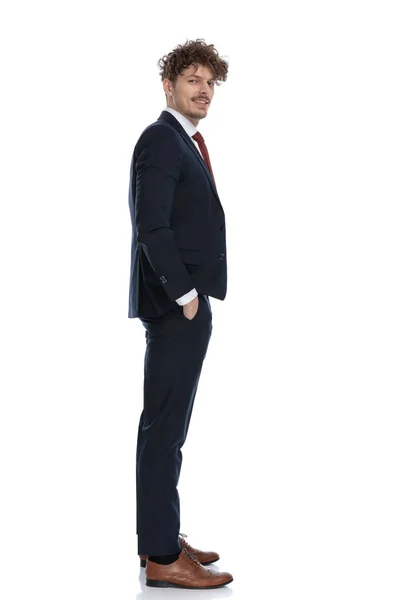 スーツを着て白いスタジオの背景に立ちながらポケットに手をつないで笑顔のビジネスマンの側面図 — ストック写真