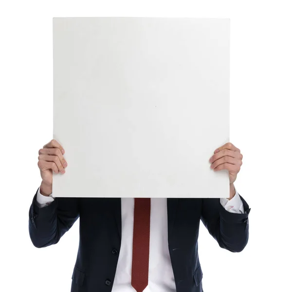 若いビジネスマンがスーツを着て白いスタジオの背景に立っている間 彼の顔を空白の看板で覆います — ストック写真
