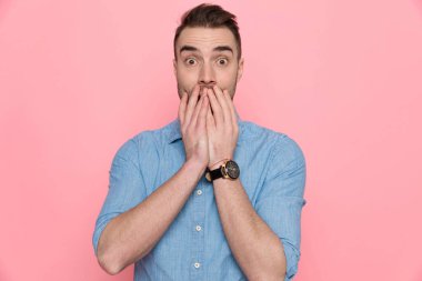 Korkmuş sıradan bir adam iki eliyle ağzını kapatıyor, mavi bir gömlek giyiyor ve pembe stüdyo arka planında duruyor.