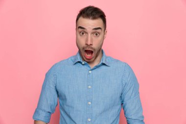 Şok olmuş sıradan bir adam ağzını kocaman açıyor, kameraya bakıyor ve pembe stüdyo arka planında mavi bir gömlek giyiyor.