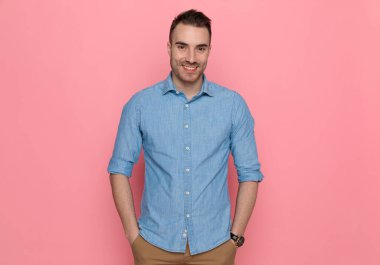 Genç, yakışıklı, sıradan bir adam elleri cebinde, gülümsüyor ve pembe stüdyo arka planında mavi bir gömlek giyiyor.