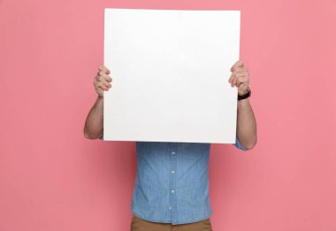 Boş bir tahtanın arkasına saklanan, mavi gömlekli ve pembe stüdyo arka planında duran sıradan bir adam.