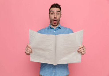 Gazeteyi tutan, ağzı açık kameraya bakan, pembe stüdyonun arka planındaki haberlerden şok olan genç ve çekici bir adam.