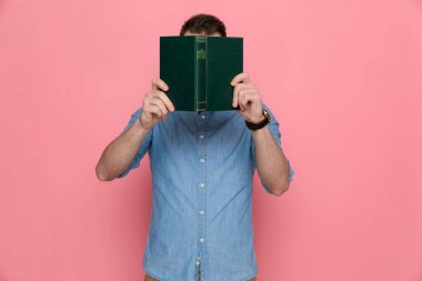 Sıradan bir adam yüzünü kitabının arkasına gizliyor, mavi bir gömlek giyiyor ve pembe stüdyo arka planında duruyor.