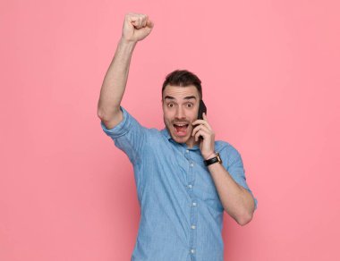 Sıradan bir adam başarıyı kutluyor, telefonla konuşuyor ve pembe stüdyo arka planında mavi bir gömlek giyiyor.