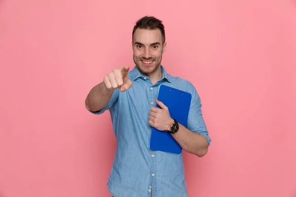 Atractivo Joven Hombre Casual Apuntando Hacia Adelante Sosteniendo Portapapeles Pecho —  Fotos de Stock