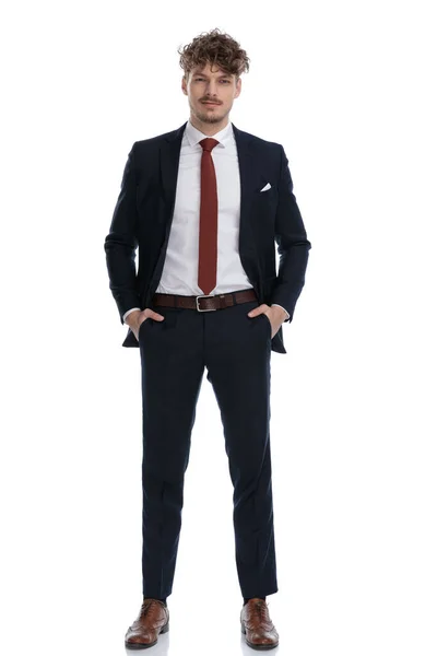 スーツを着て白いスタジオの背景に立っている間 彼のポケットに両手で楽しみにしている自信のビジネスマン — ストック写真