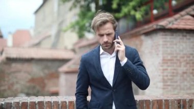 Zarif bir iş adamı bir eli cebinde tuğla bir duvara dayamış telefonla konuşuyor ve etrafa bakıyor.