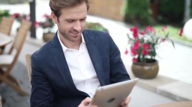 Sarışın yakışıklı iş adamı tabletine mesaj atıyor, masada oturuyor, gülümsüyor ve başparmağını kaldırıyor.
