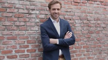 Kolları göğsünde çaprazlanmış, tuğladan duvara yaslanmış ve gülümseyen, etrafa bakınan genç ve zeki iş adamı.