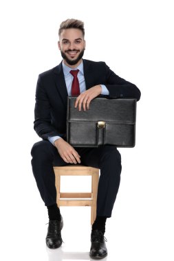 Takım elbiseli mutlu genç işadamı gülümsüyor ve elinde bavulla beyaz arka planda tek başına oturuyor.