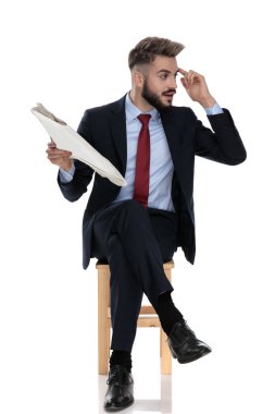 Genç işadamı gazeteyi tutuyor ve parmağıyla işaret ediyor, beyaz arka planda tek başına oturuyor.