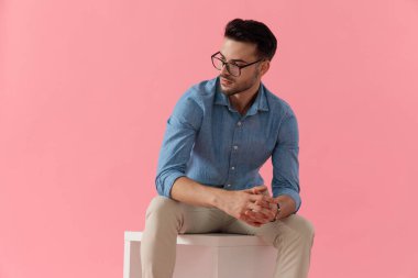 Gözlük takan çekici bir iş adamı elleri kucağında oturmuş pembe stüdyo arka planında dramatik bakışlar atıyor.