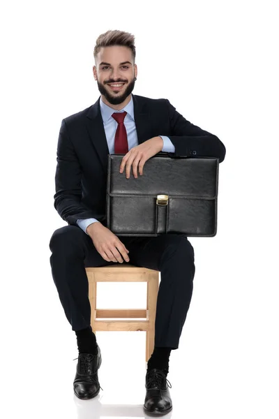 Feliz Joven Hombre Negocios Traje Sonriendo Sosteniendo Maleta Sentado Aislado — Foto de Stock