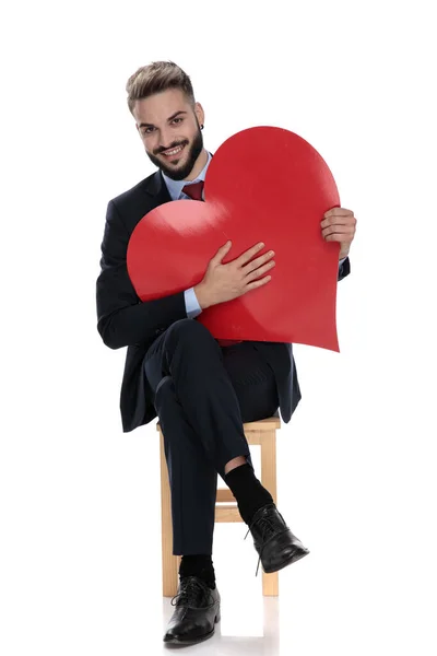 Sorridente Jovem Empresário Segurando Grande Coração Vermelho Sentado Isolado Fundo — Fotografia de Stock