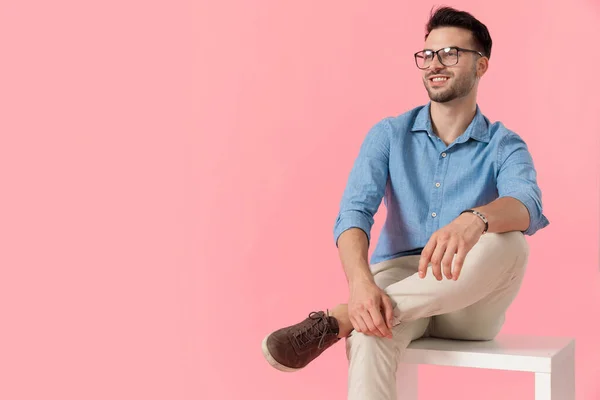 attractive businessman wearing eyeglasses sitting on cube with leg resting on the other while looking away happy on pink studio background