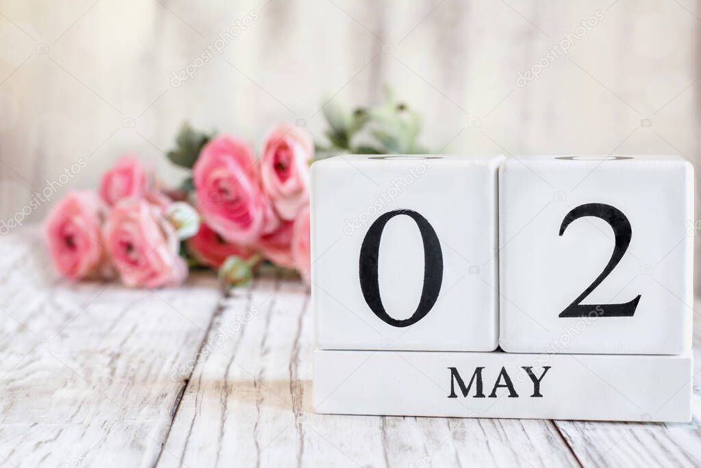 White wood calendar blocks with the date May 2nd. Selective focus with pink ranunculus in the background over a wooden table.