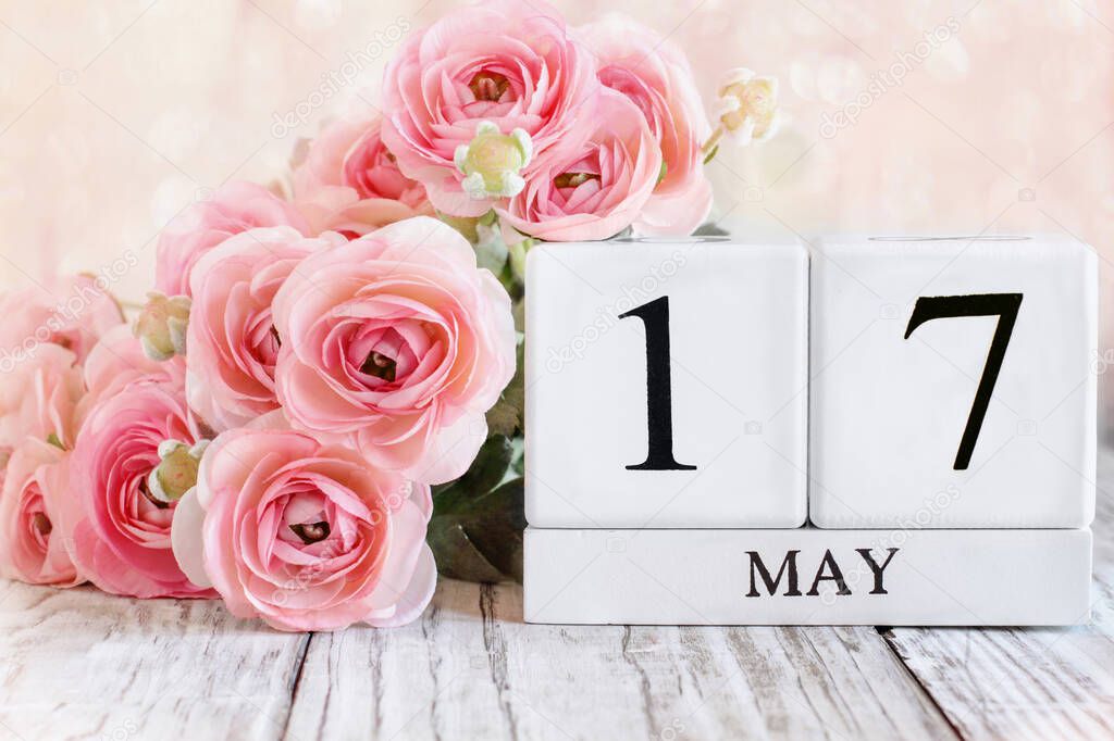 White wood calendar blocks with the date May 17th and pink ranunculus flowers over a wooden table. Selective focus with blurred background. 