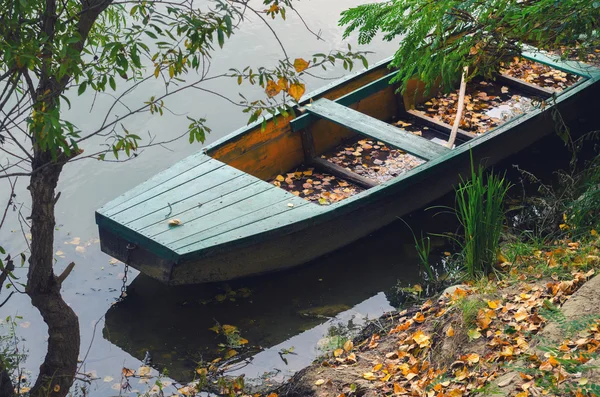 Деревянная рыбацкая лодка на берегу реки, покрытая опавшими листьями осенью — стоковое фото
