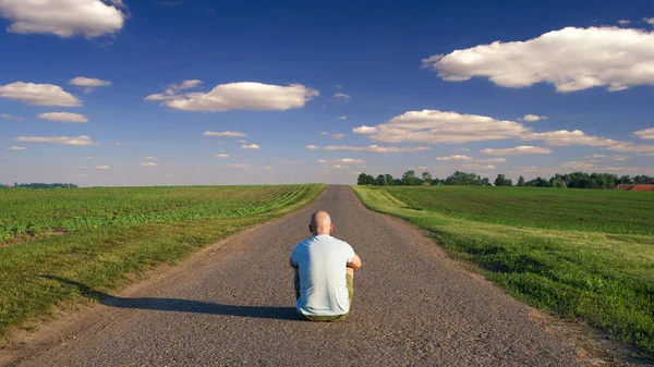 Hombre sentado en el medio de la carretera del campo en el soleado día de verano —  Fotos de Stock