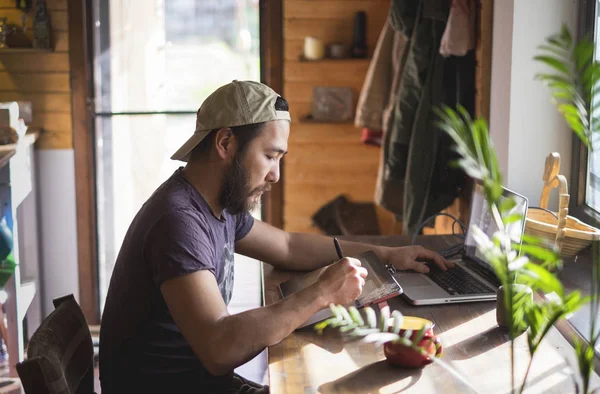 asian man with beard working on a tablet and a laptop at home